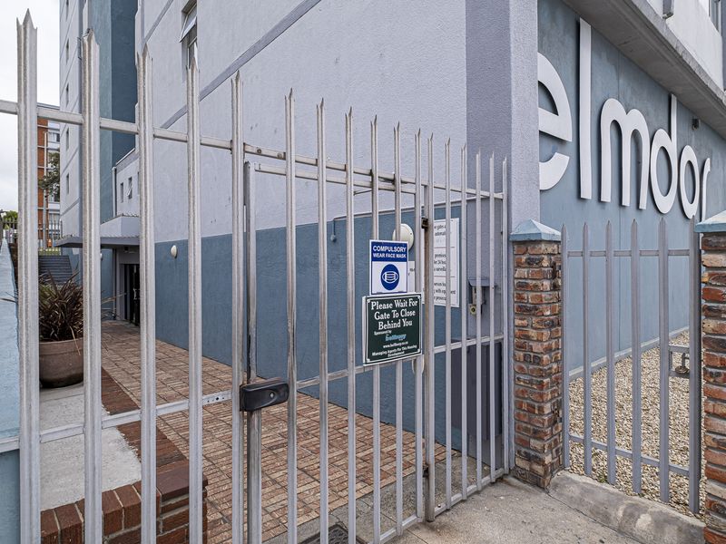 Exterior view of the apartment building entrance featuring a secure metal gate, brick pillars, and paved access path.