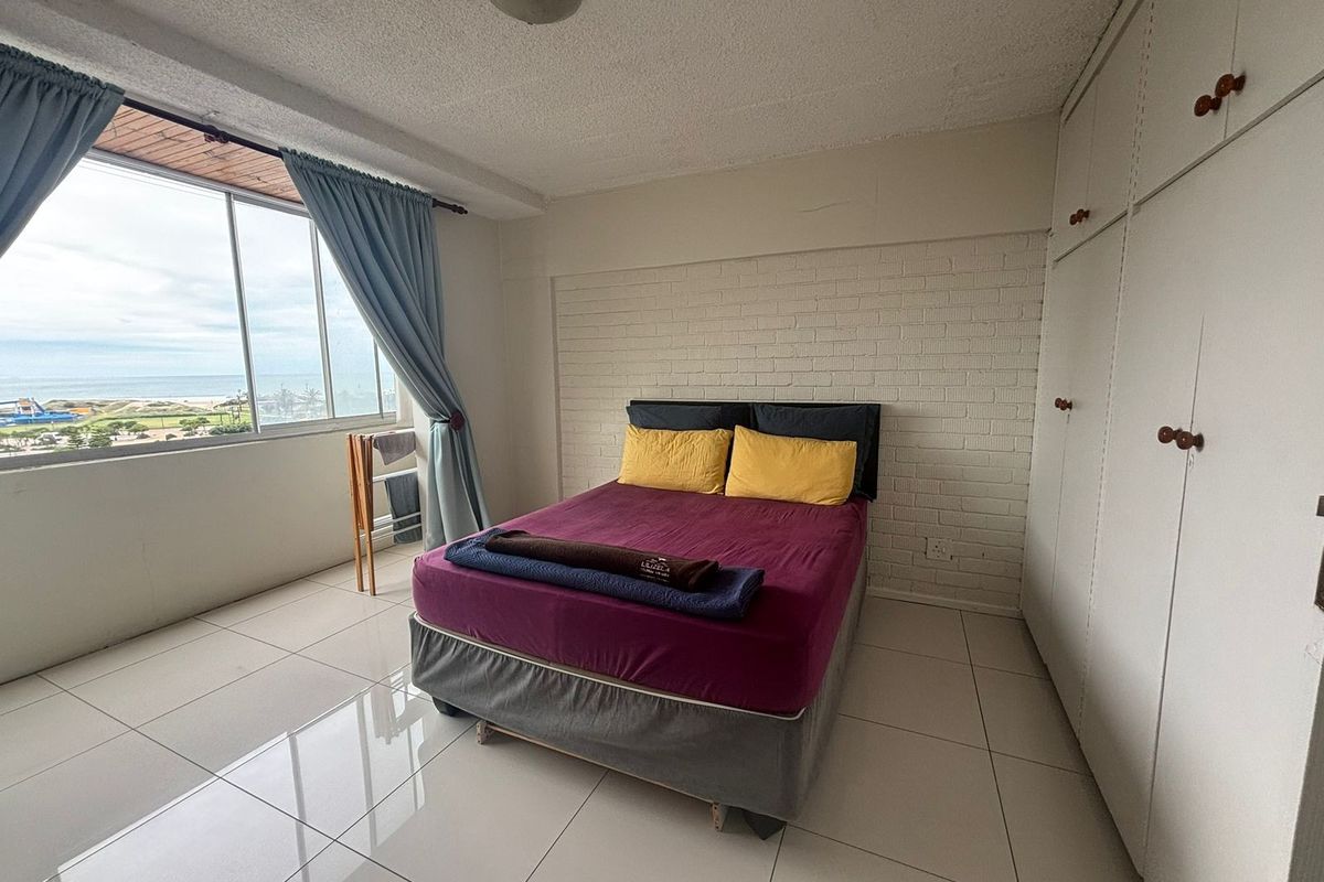 A bedroom featuring tiled floors, a painted brick accent wall, and built-in cupboards, offering natural light and a view through a large window.