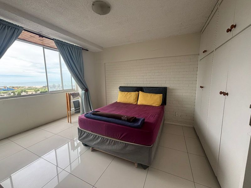 A bedroom featuring tiled floors, a painted brick accent wall, and built-in cupboards, offering natural light and a view through a large window.