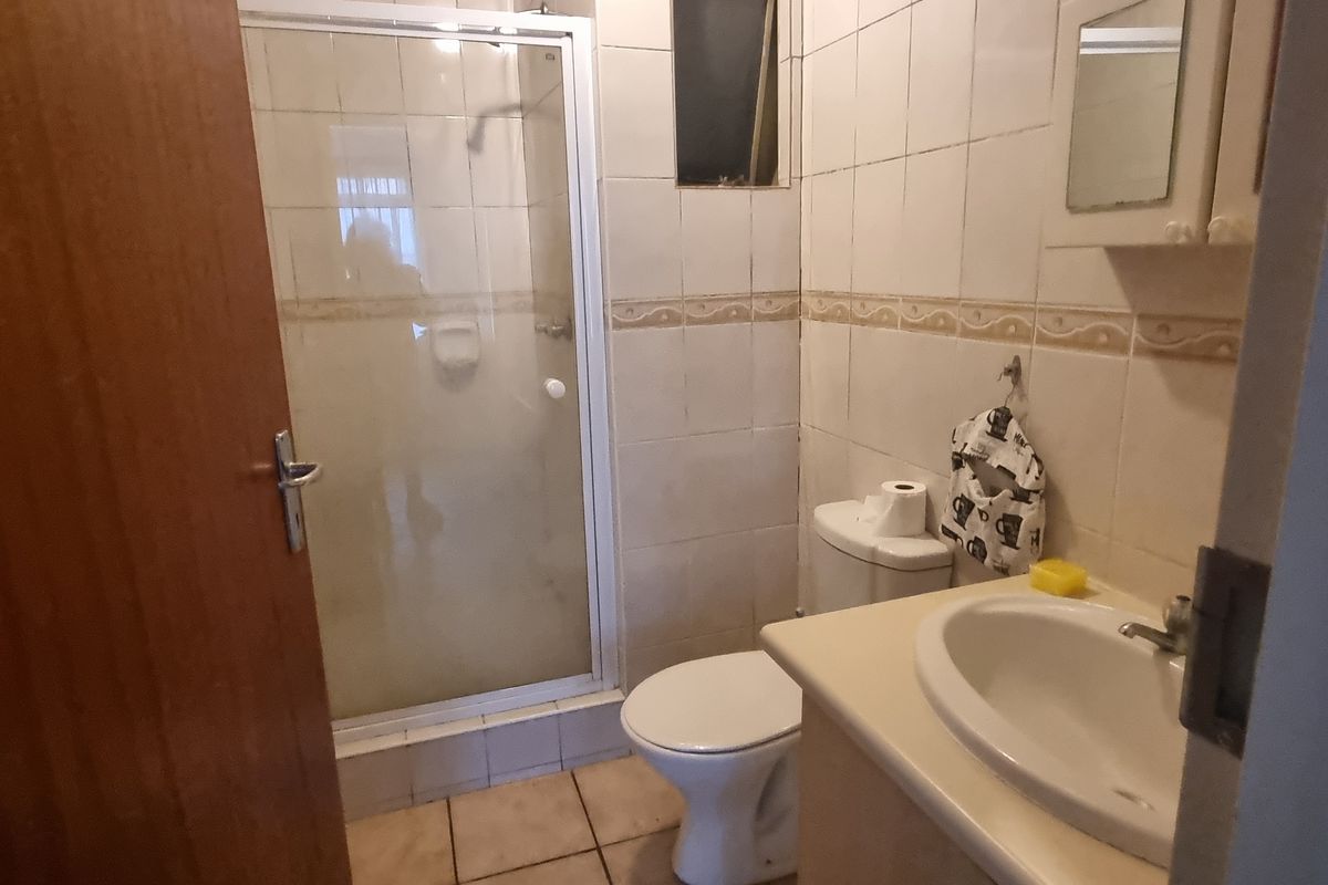 A compact bathroom featuring a shower enclosure, toilet, and vanity basin with tiled walls and flooring, presenting a dated aesthetic.