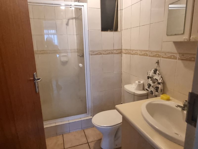 A compact bathroom featuring a shower enclosure, toilet, and vanity basin with tiled walls and flooring, presenting a dated aesthetic.