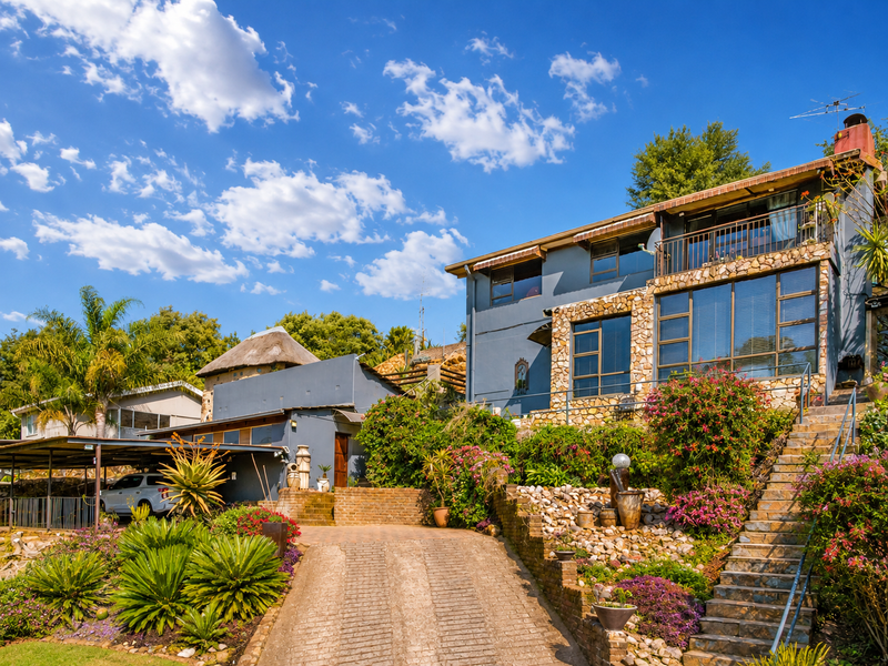 Front elevation of a multi-level residence featuring stone cladding, a paved driveway, a carport, and landscaped terraced gardens with stone retaining walls.