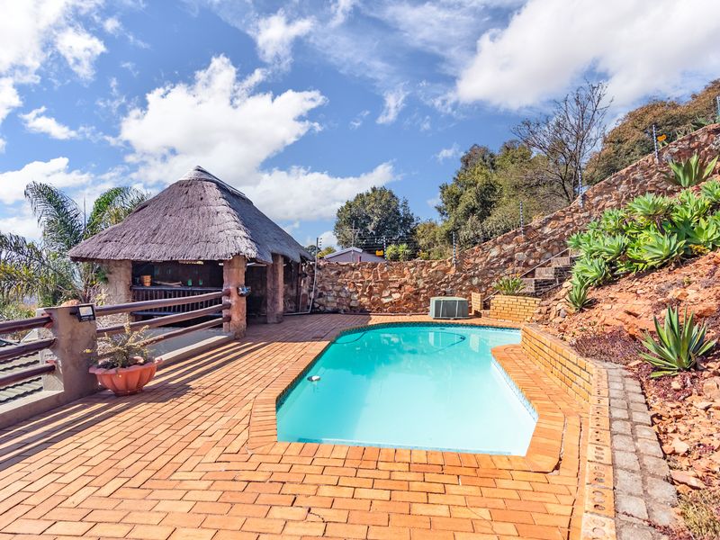 Outdoor pool area featuring a swimming pool, brick paved surround, and a thatched lapa set against a natural stone retaining wall.