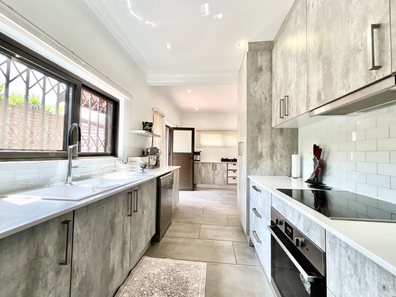 Modern galley-style kitchen featuring concrete-look cabinetry, subway tile backsplash, integrated oven, electric cooktop, and sleek tiled flooring with ample natural light.