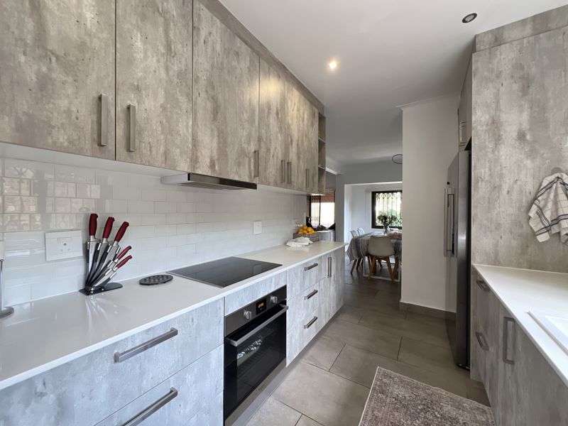 Modern galley-style kitchen featuring concrete-look cabinetry, white quartz countertops, subway tile backsplash, and integrated appliances, leading into an adjacent dining area.