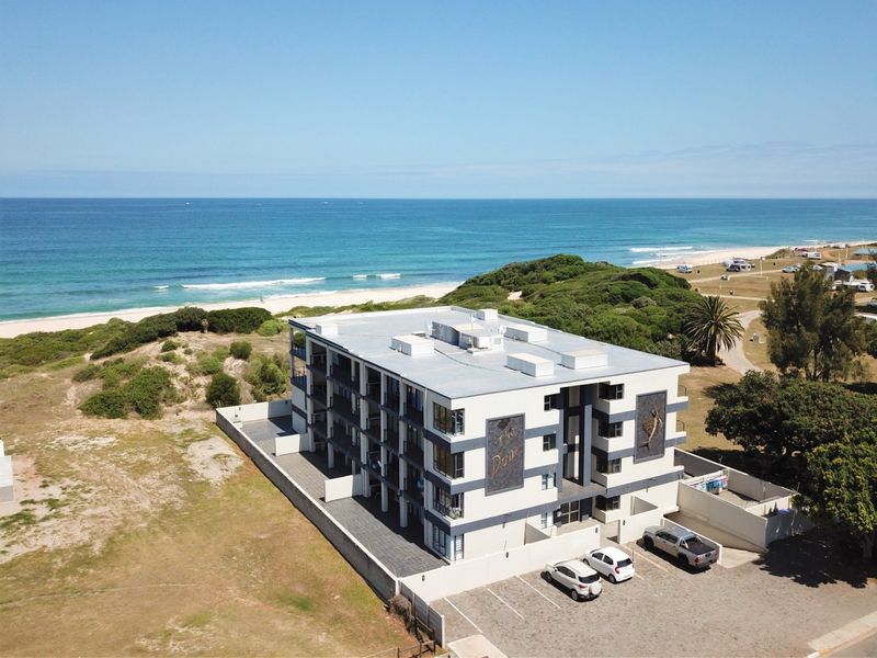 Aerial view of The Dune apartment complex, showcasing its proximity to the coastline, paved parking facilities, and multi-story building structure.