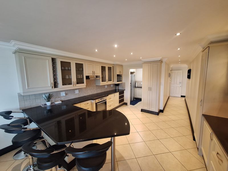 A well-maintained kitchen featuring cream cabinetry, black granite countertops, a breakfast bar, and tiled flooring, with an adjacent scullery area.