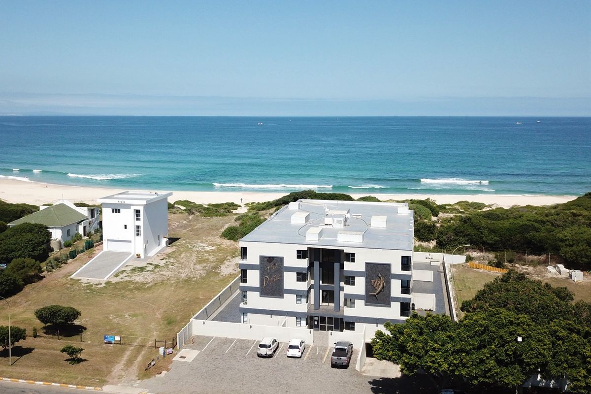 Aerial view of a modern apartment complex situated directly on the beachfront, featuring a paved parking area and secure perimeter wall.