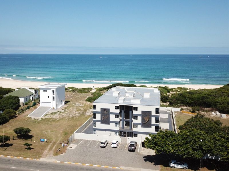 Aerial view of a modern apartment complex situated directly on the beachfront, featuring a paved parking area and secure perimeter wall.