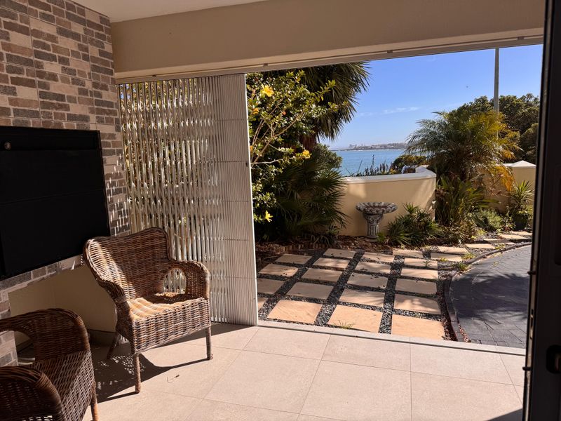 Covered patio featuring a built-in fireplace, brick feature wall, and tiled flooring, opening onto a landscaped garden with scenic coastal views.