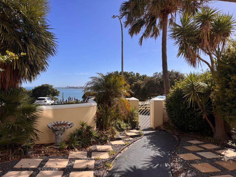A well-maintained front entrance area featuring a paved walkway, landscaped garden, and a pedestrian gate with a scenic coastal view.