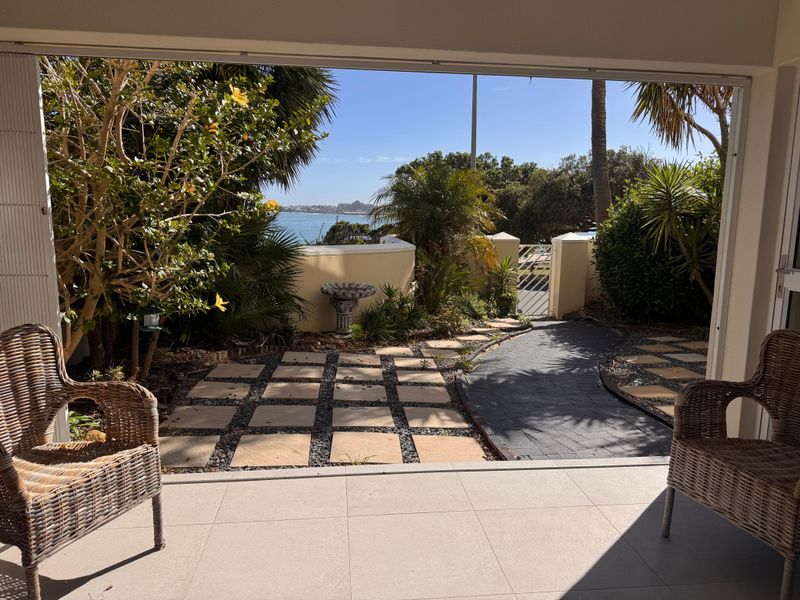 A covered patio area featuring tiled flooring, overlooking a landscaped garden with a paved path and scenic ocean views.