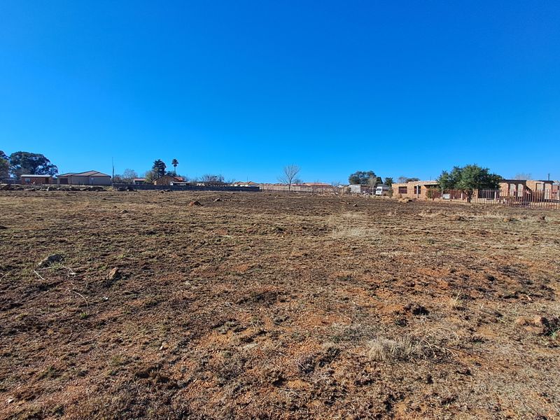 A wide-angle view of a vacant residential plot of land with natural ground cover and perimeter fencing visible in the distance.
