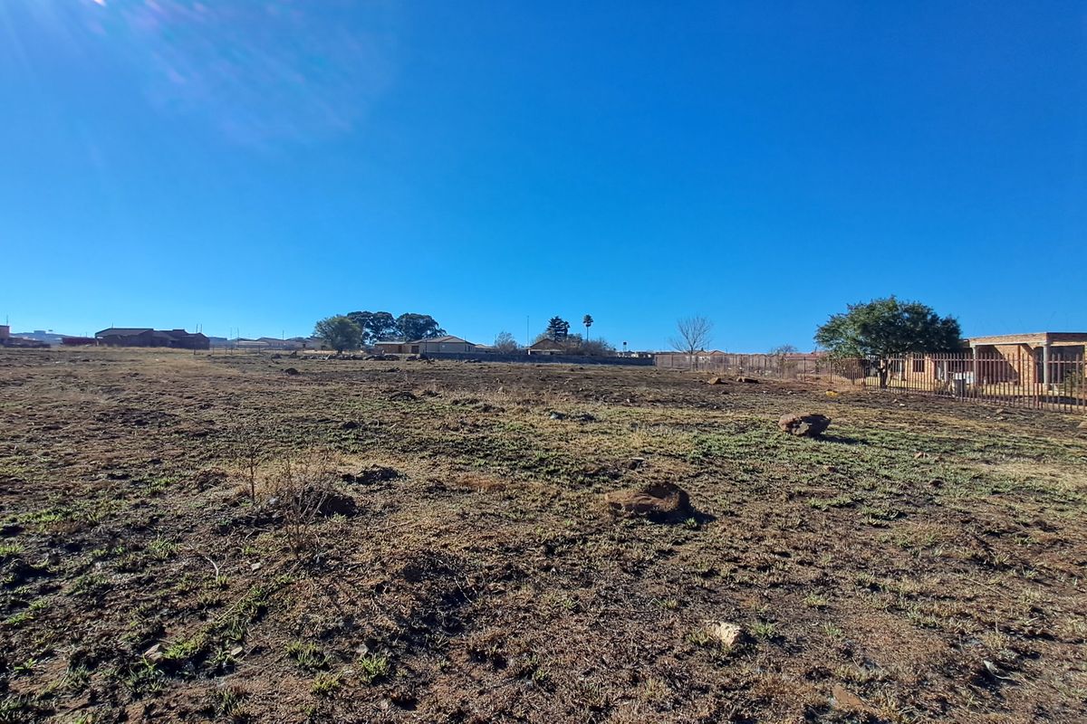 A wide-angle view of a vacant residential plot featuring dry ground and perimeter fencing, situated in a developed neighborhood.