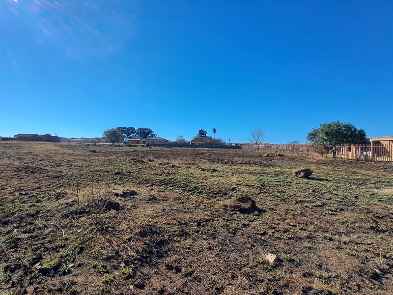 A wide-angle view of a vacant residential plot featuring dry ground and perimeter fencing, situated in a developed neighborhood.