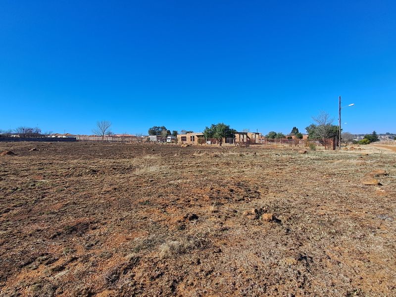 A wide-angle view of a vacant residential plot featuring natural terrain, perimeter fencing, and surrounding neighborhood infrastructure.