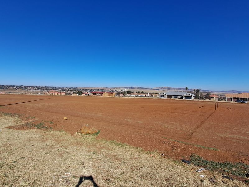 A wide-angle view of a large, cleared vacant residential plot with surrounding neighborhood houses visible in the distance.