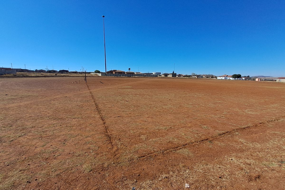 A wide-angle view of a vacant residential plot featuring dry earth and a tall floodlight pole under a clear blue sky.