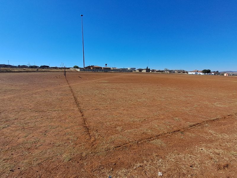 A wide-angle view of a vacant residential plot featuring dry earth and a tall floodlight pole under a clear blue sky.