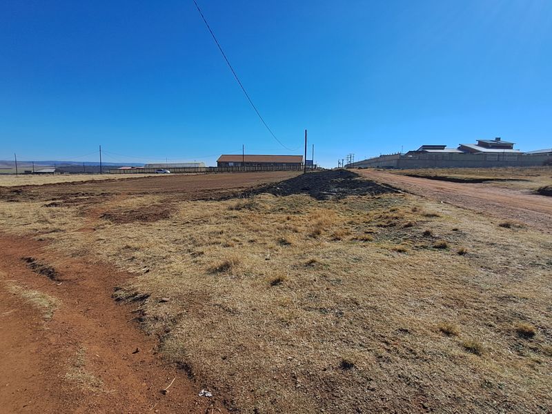 A wide-angle view of a vacant residential plot featuring natural terrain, adjacent dirt road access, and surrounding neighborhood boundary walls.