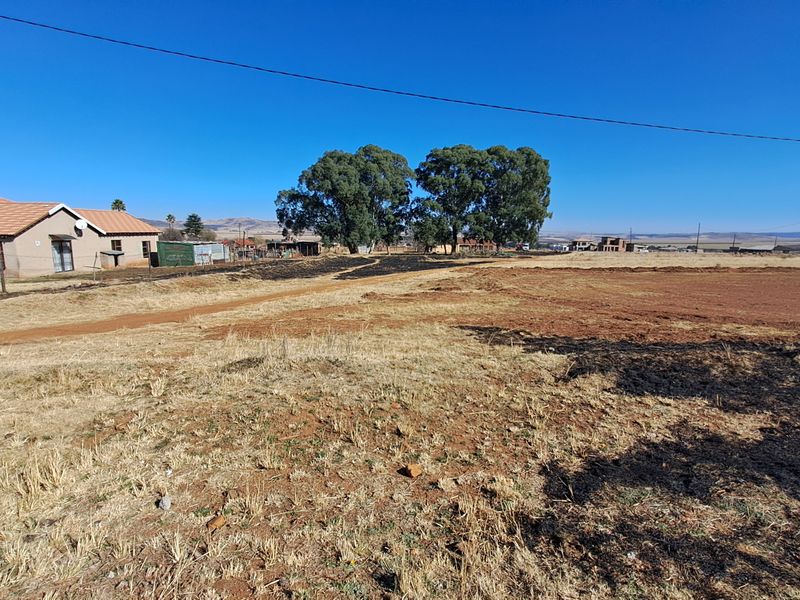 A wide-angle view of a vacant residential plot featuring dry, unmaintained land with a dirt access path and surrounding rural landscape.