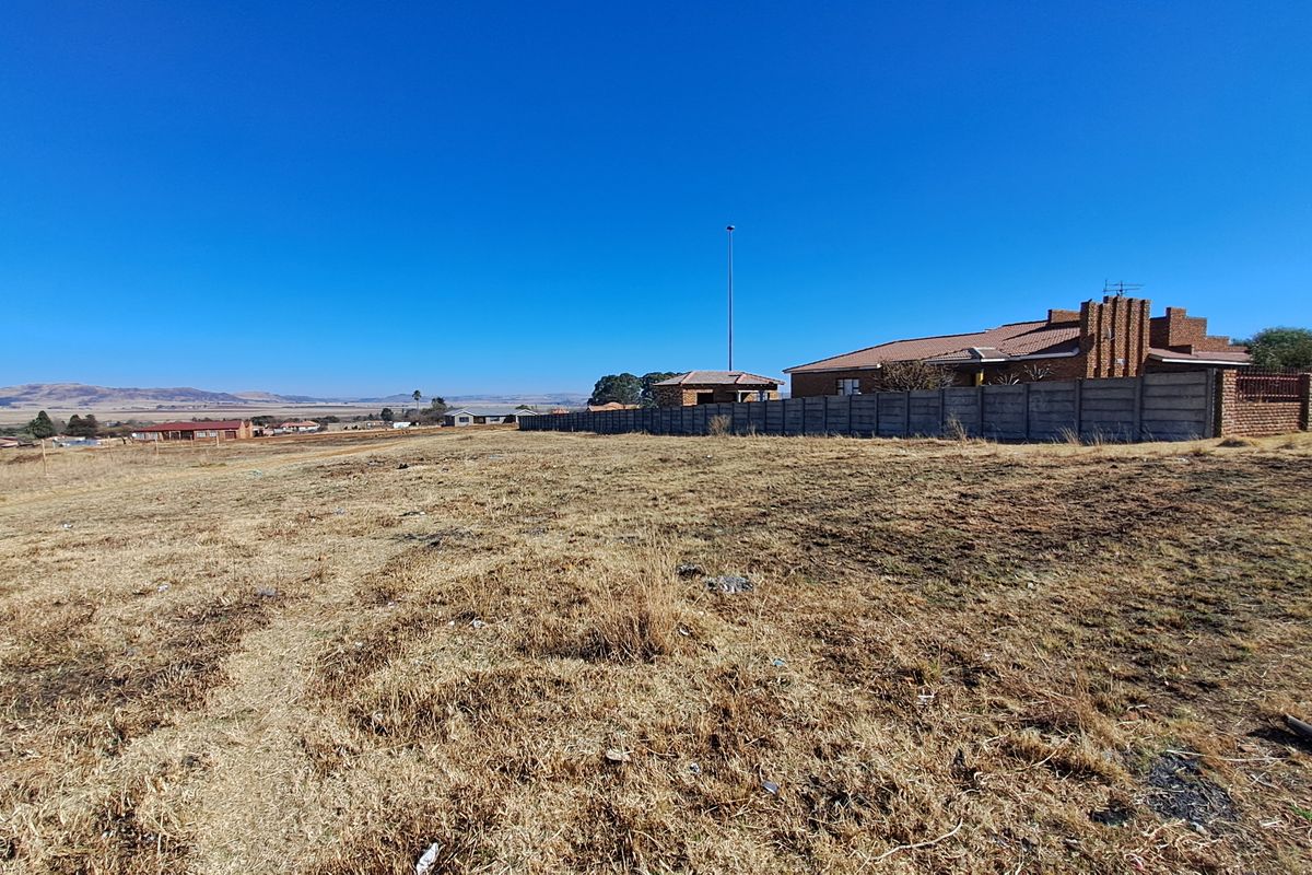 A wide-angle view of a vacant residential plot featuring dry grass, a precast concrete boundary wall, and distant neighborhood views.