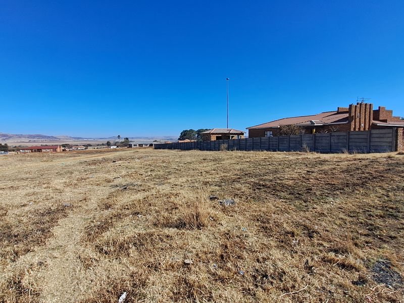 A wide-angle view of a vacant residential plot featuring dry grass, a precast concrete boundary wall, and distant neighborhood views.