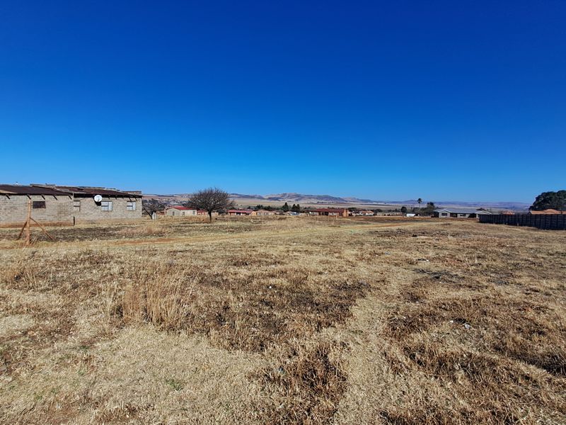 A wide-angle view of a vacant residential plot featuring dry grass, perimeter fencing, and an adjacent masonry structure in need of repair.
