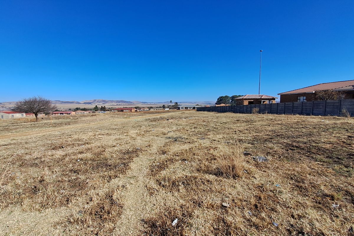 A wide-angle view of a vacant residential plot featuring dry grass, a precast concrete boundary wall, and distant neighborhood landscape.