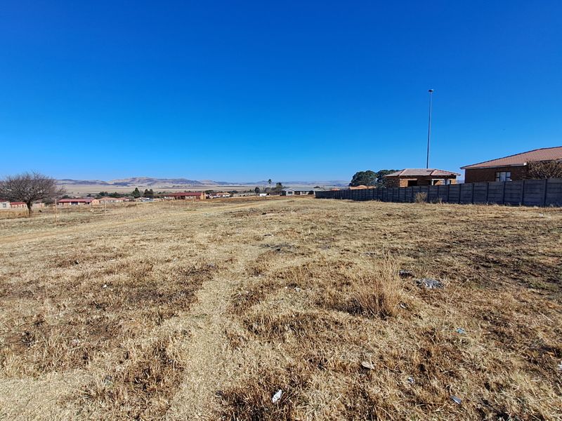 A wide-angle view of a vacant residential plot featuring dry grass, a precast concrete boundary wall, and distant neighborhood landscape.