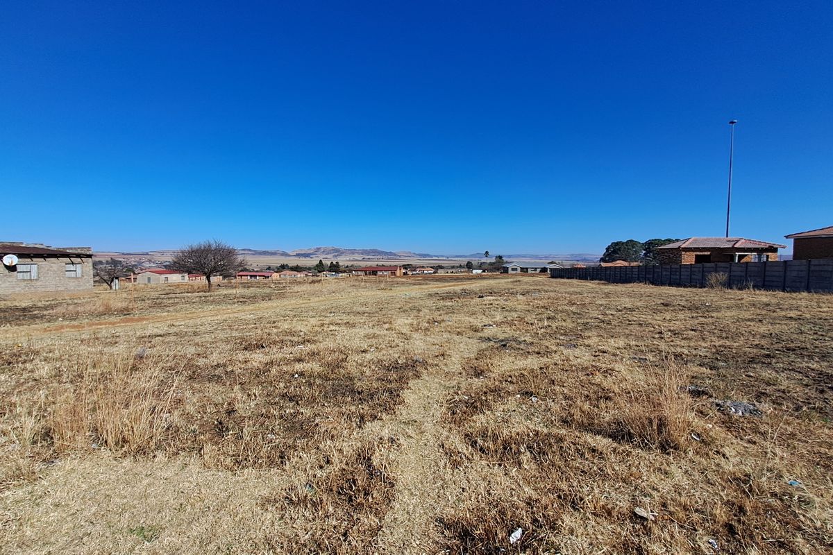 A wide-angle view of a vacant residential plot featuring dry grass, a precast concrete boundary wall, and surrounding neighborhood structures.