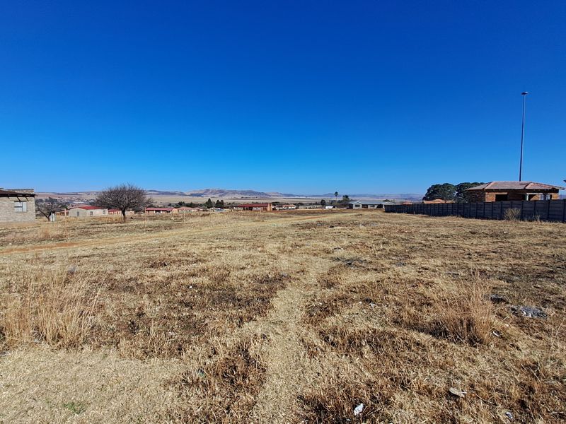 A wide-angle view of a vacant residential plot featuring dry grass, a precast concrete boundary wall, and surrounding neighborhood structures.