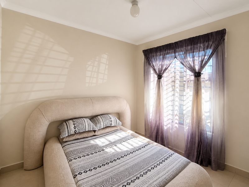 A well-lit bedroom featuring neutral wall tones, tiled flooring, and a window fitted with security bars and decorative curtains.