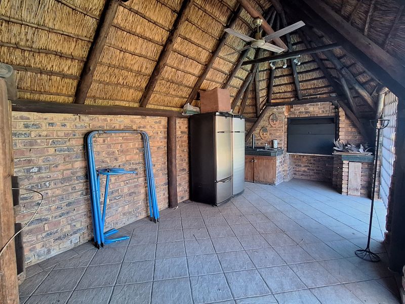 Covered outdoor patio featuring a traditional thatched roof, exposed brickwork, built-in braai, and a functional kitchenette area with sink and cabinetry.