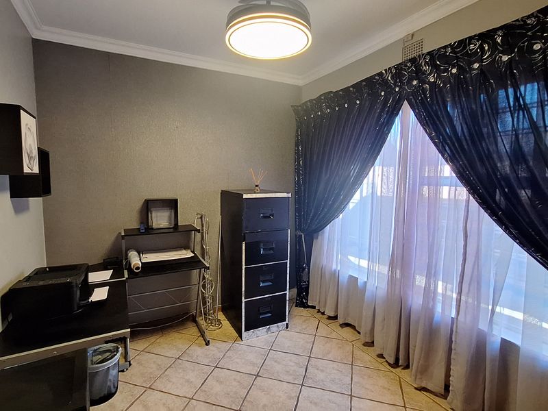A functional home office space featuring tiled flooring, a central ceiling light fixture, and a large window with decorative curtains.