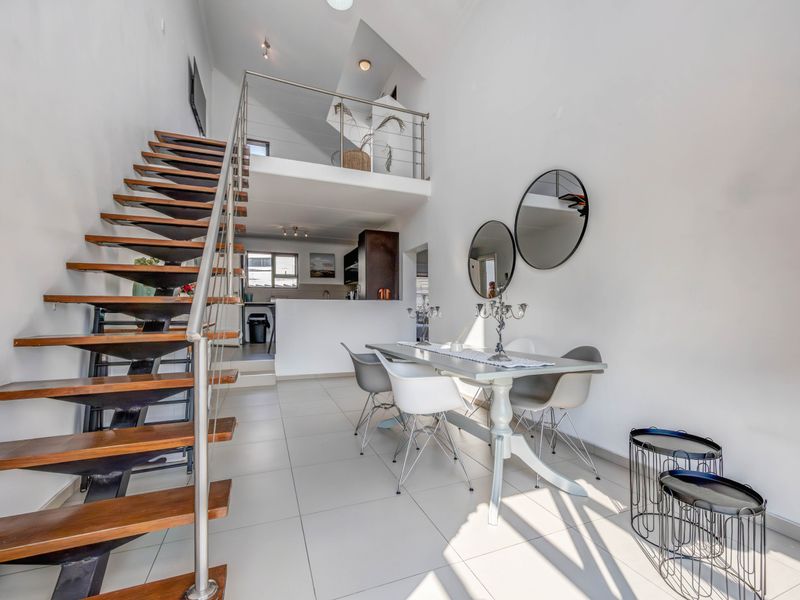 Open plan living and dining area featuring a modern staircase with stainless steel balustrades leading to a mezzanine level, finished with neutral floor tiles.