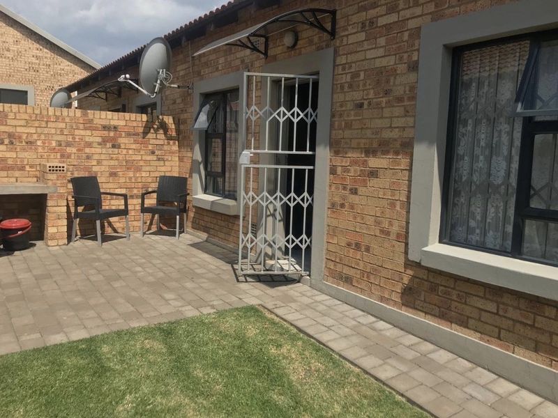 Rear exterior view of the apartment featuring a paved patio area, built-in brick braai, small lawn, and secure entrance with a metal security gate.