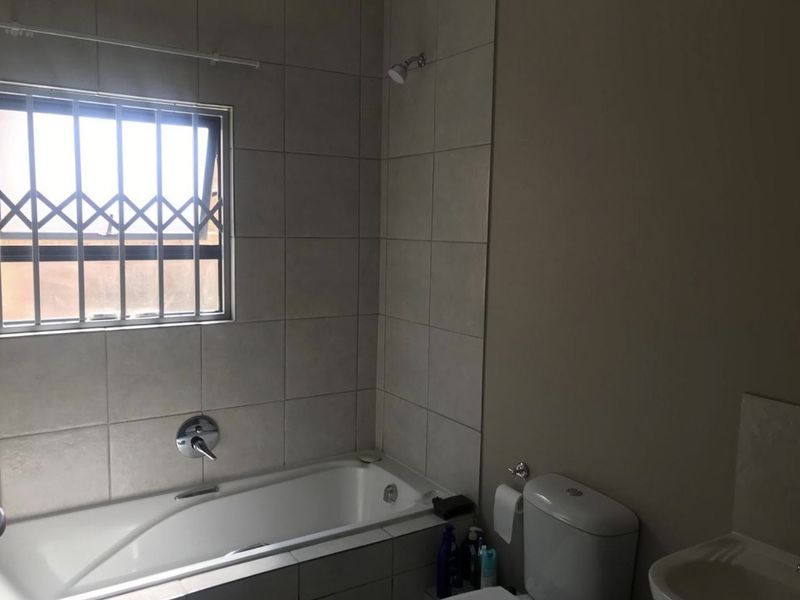 A functional bathroom featuring a bathtub with shower head, toilet, and basin, finished with neutral wall tiling and a window with security bars.