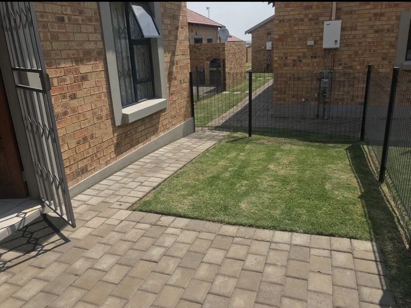 A private rear garden area featuring a paved patio, small lawn, and secure perimeter fencing, adjacent to the face brick apartment exterior.