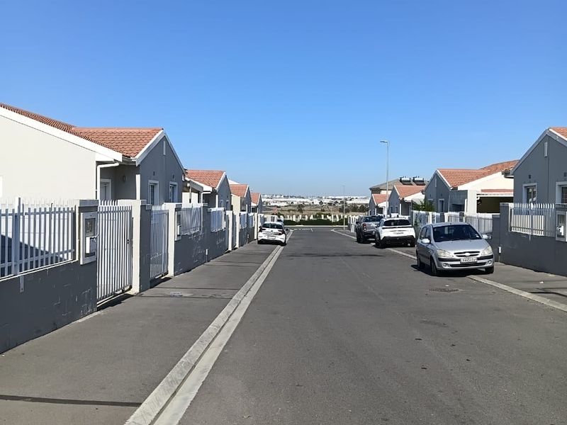 A view of a residential street featuring uniform houses with tiled roofs and metal perimeter fencing along an asphalt road.