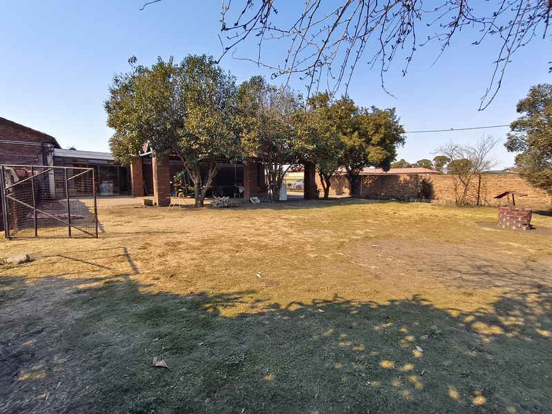 A spacious backyard featuring a large lawn, mature trees, a covered patio area, and a brick fire pit, enclosed by a perimeter wall.