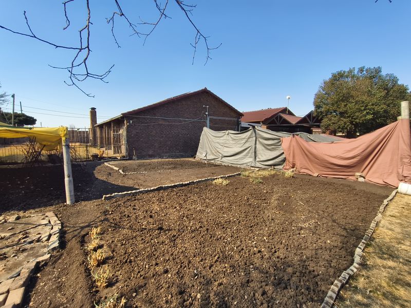 A view of a residential garden area featuring exposed soil beds, brick building structure, and temporary makeshift shade coverings.