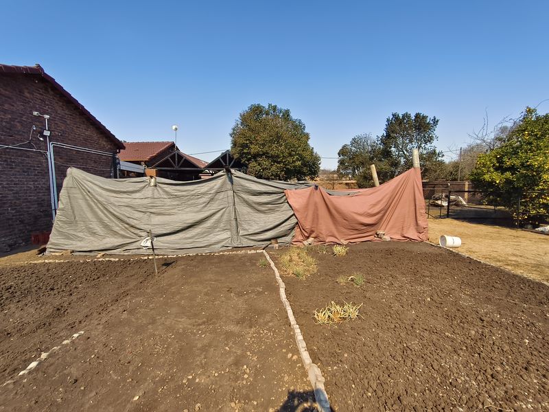 A residential garden area featuring tilled soil beds, brick edging, and temporary tarp screening against the boundary wall.