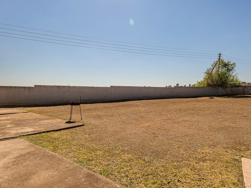 A spacious, open backyard featuring a concrete patio area and a large, dry lawn enclosed by a perimeter boundary wall.