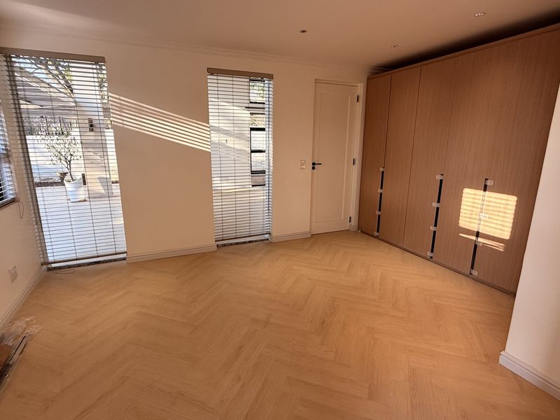 A bright, empty bedroom featuring modern herringbone timber flooring, built-in wooden cupboards, and large windows with fitted venetian blinds.