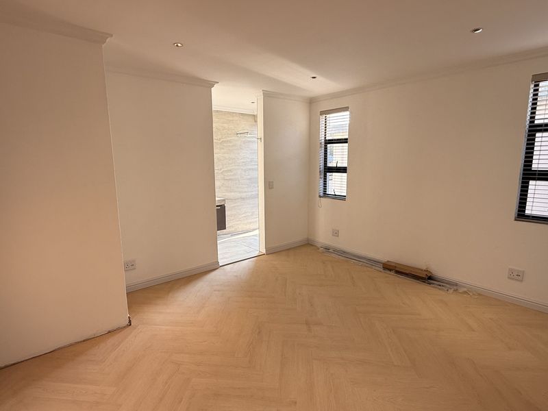 A bright, empty bedroom featuring herringbone wood flooring, recessed lighting, and an adjoining en-suite bathroom, ready for personalization.