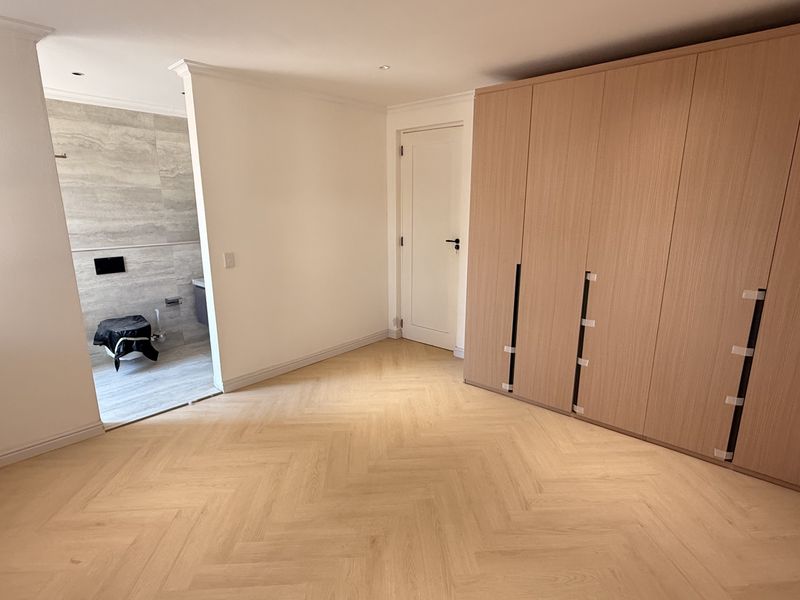 A bright, modern bedroom featuring light wood herringbone flooring, extensive built-in cabinetry, and an open-plan transition to an en-suite bathroom.