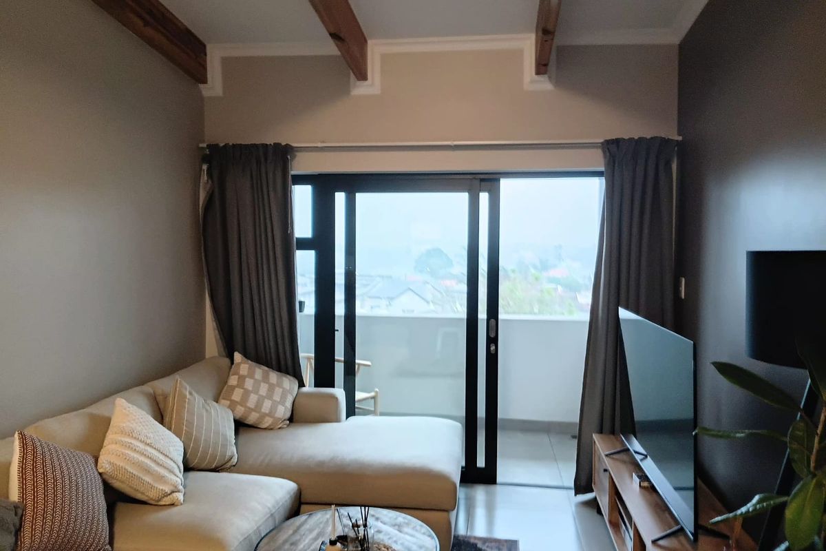 A well-maintained living room featuring exposed timber ceiling beams, tiled flooring, and large sliding glass doors providing access to a private balcony.