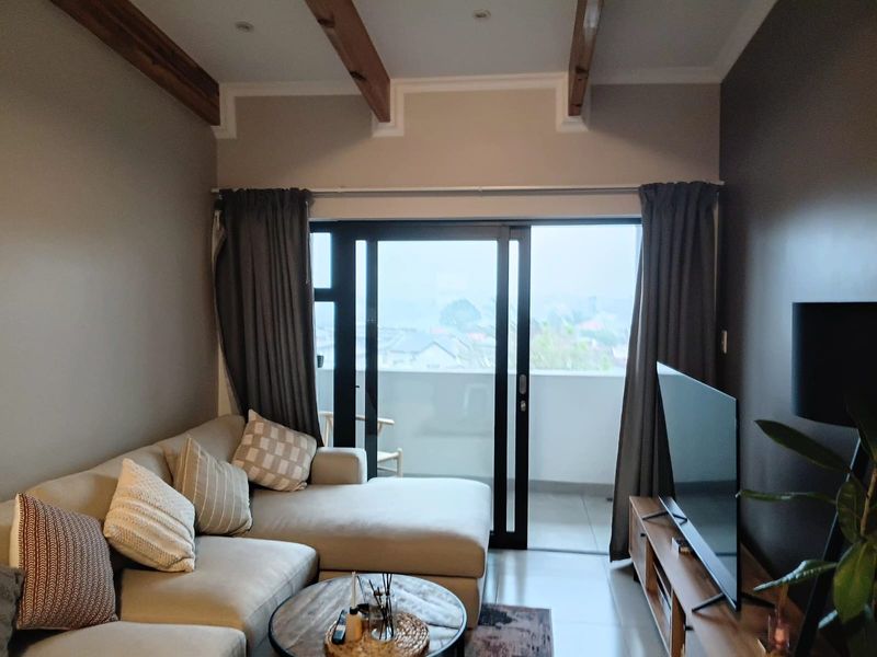 A well-maintained living room featuring exposed timber ceiling beams, tiled flooring, and large sliding glass doors providing access to a private balcony.