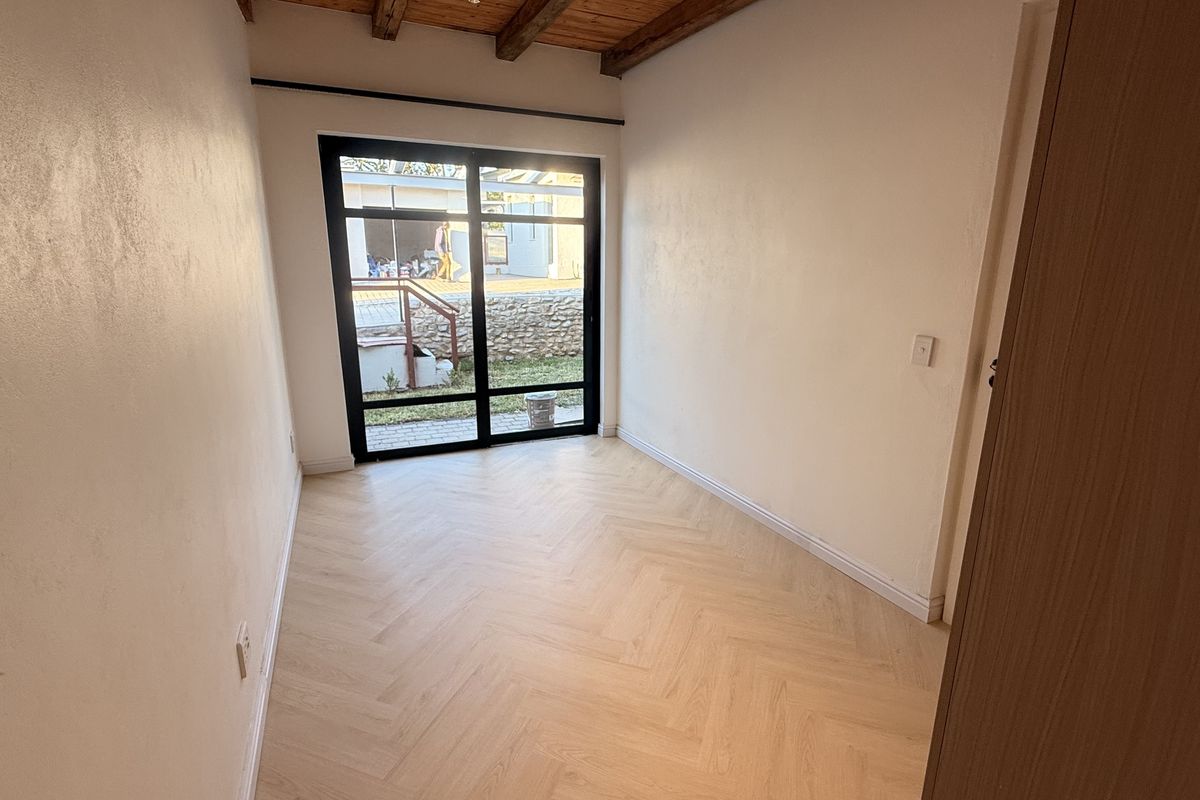 A bright, empty bedroom featuring modern herringbone laminate flooring, exposed timber ceiling beams, and a large black-framed glass sliding door leading to the exterior.